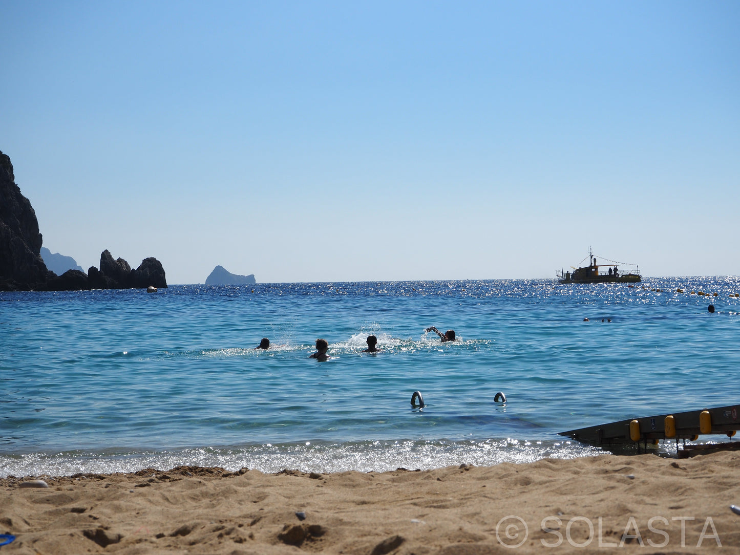 People swimming in blue ocean water near a sandy beach with a boat anchored in the distance
