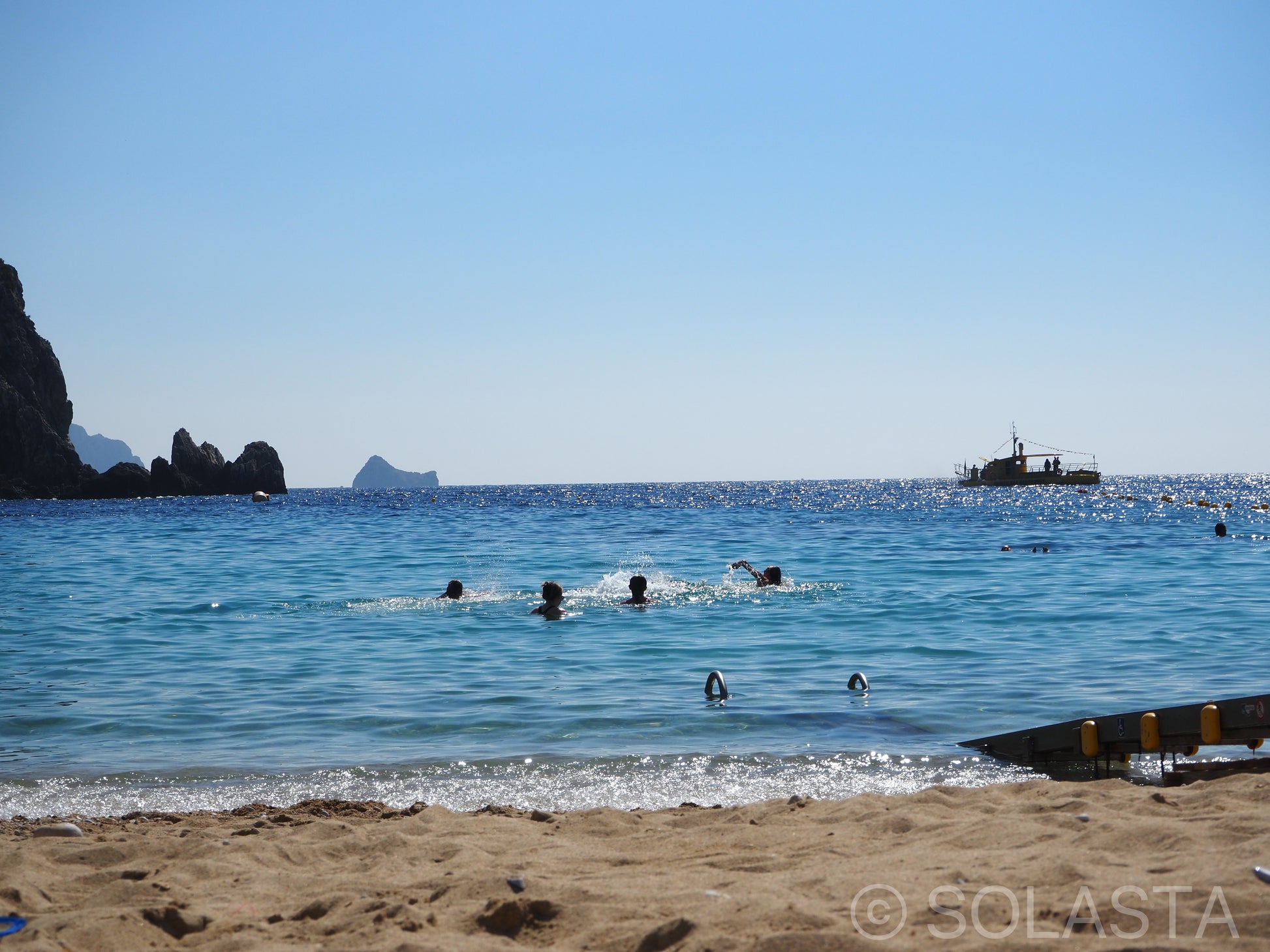 People swimming in blue ocean water near a sandy beach with a boat anchored in the distance